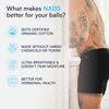 Tattooed man wearing black NADS boxer briefs in a kitchen, with overlay text highlighting organic cotton, no harsh chemicals, breathable fabric, and hormonal health benefits.